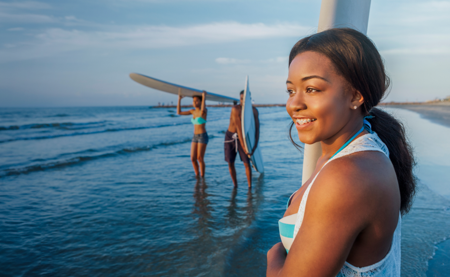 A smiling young woman standing on a beach holding a surfboard upright while looking out at the water and two friends are behind her carrying their surfboards