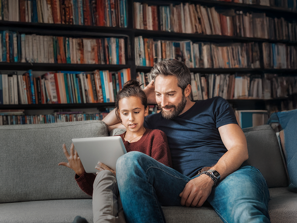 A father and his young daughter sitting on a couch and looking at a tablet