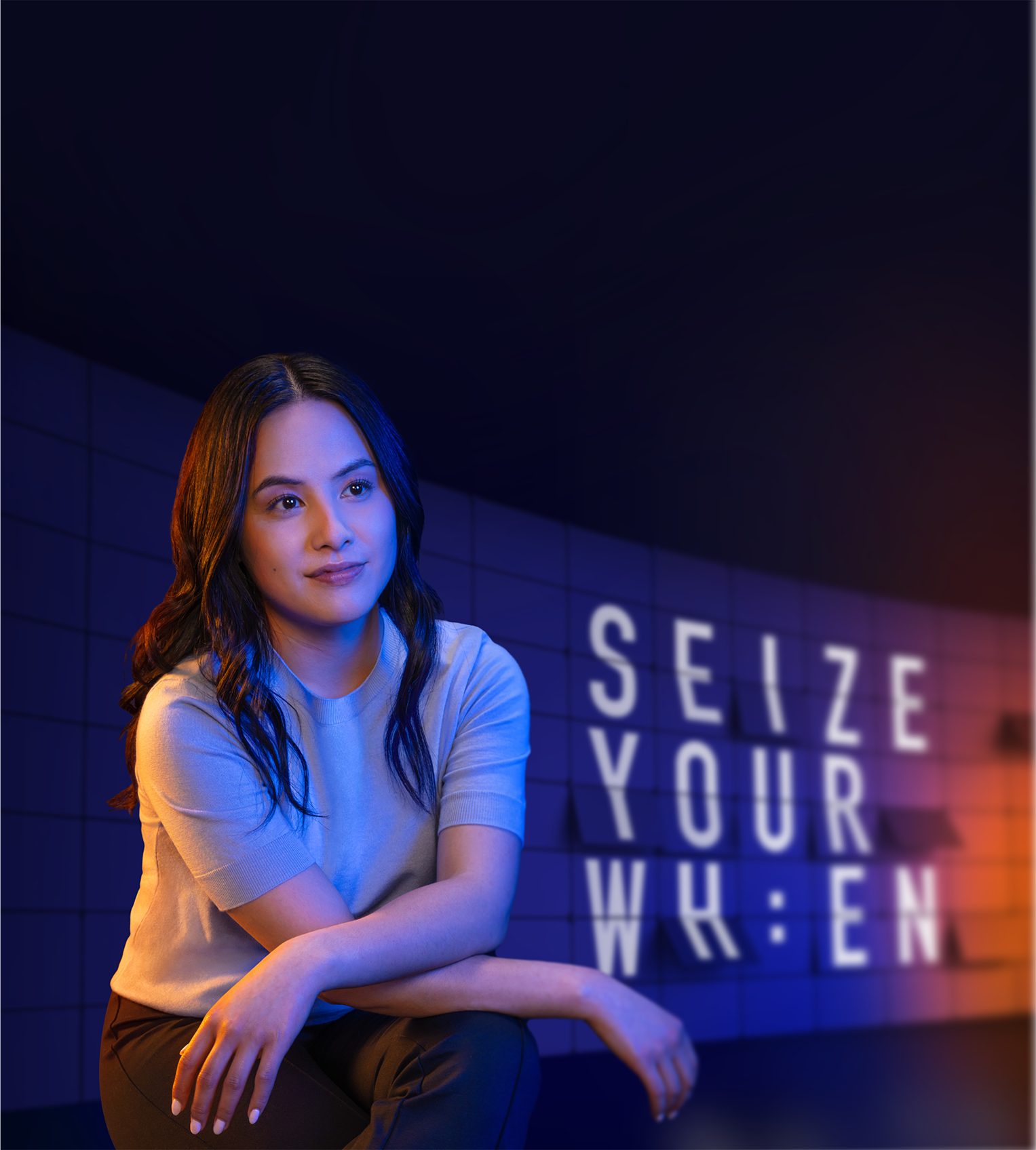 A woman posing with her arms crossed on her knee in front of a backdrop that reads "SEIZE YOUR WHEN"